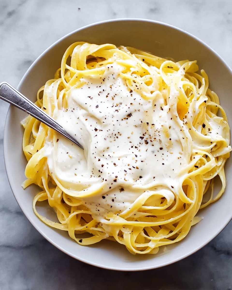 A bowl with one layer of yellow pasta noodles arranged loosely in a circular shape, topped with a smooth, creamy white sauce evenly spread over the noodles. The sauce has a soft texture with small black pepper flakes sprinkled on top. The bowl is white and sits on a white marbled surface. A silver fork rests inside the bowl partly on the pasta. Photo taken with an iphone --ar 4:5 --v 7
