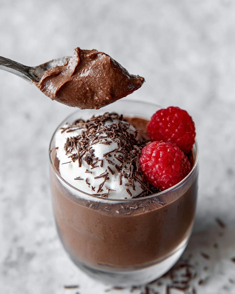 A clear glass cup shows three visible layers: the base is a smooth, dark brown chocolate mousse filling most of the cup, the middle has a small dollop of white whipped cream slightly off-center, and two bright red raspberries rest next to the cream on the top right. Dark chocolate shavings are sprinkled over the cream and mousse. A spoon with a silver handle holds a scoop of the chocolate mousse mixed with some whipped cream and chocolate shavings, hovering just above the cup. The background is a white marbled texture. Photo taken with an iphone --ar 4:5 --v 7