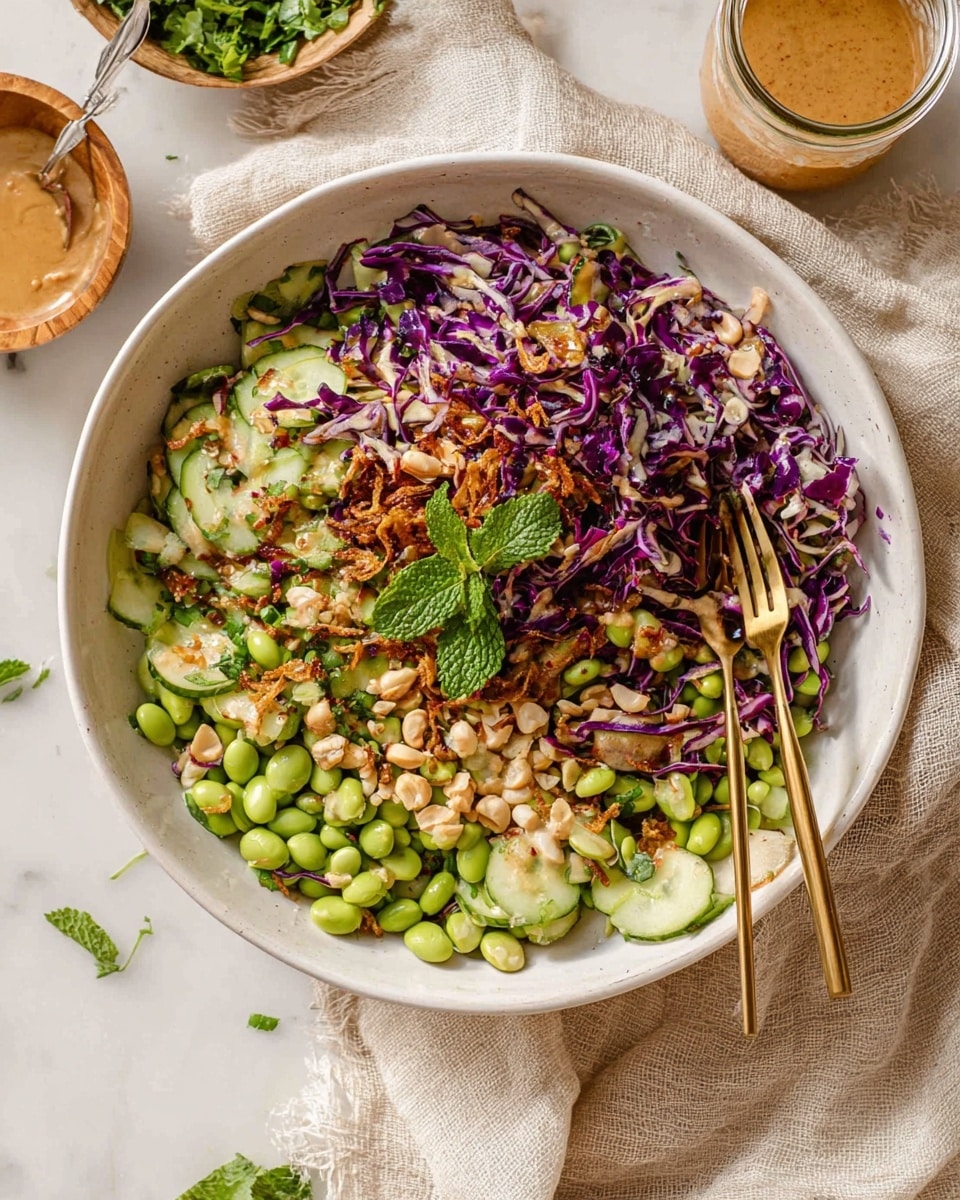 A large white bowl filled with a colorful salad showing three main layers: thin, curled purple cabbage shreddings on top, scattered pale green edamame beans and sliced round cucumber pieces spread evenly in the middle, and light brown chopped nuts mixed throughout. Golden fried shallots are sprinkled on top, adding texture. Near the center, a few fresh green mint leaves sit as a garnish. A pair of golden chopsticks hold some of the salad on the right side. The bowl rests on a light cloth on a white marbled surface. In the background, there is a glass jar with dressing and a small wooden bowl with creamy sauce visible, along with some green herbs around. Photo taken with an iphone --ar 4:5 --v 7