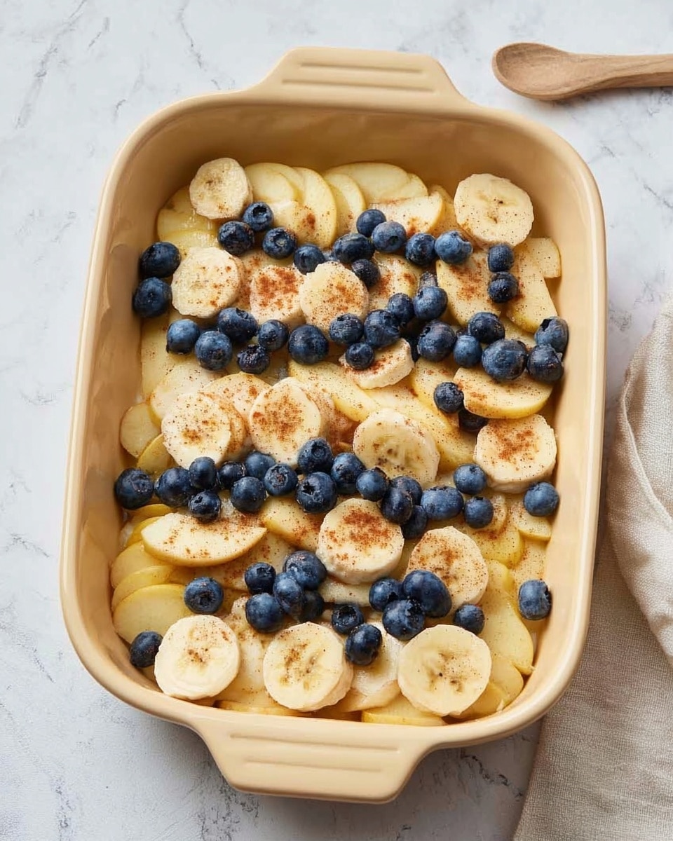 A tan rectangular baking dish filled with three layers: the bottom layer consists of sliced light yellow apples spread evenly, the middle layer has sliced bananas placed on top, and the top layer is scattered with whole blueberries and a light sprinkle of brown spices. The dish is set on a white marbled textured surface with a light-colored cloth partially visible on the bottom right side. photo taken with an iphone --ar 4:5 --v 7