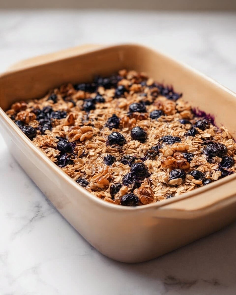 A rectangular beige baking dish filled with a baked oat and blueberry dish, showing one even layer of golden brown oats mixed with scattered dark blue blueberries throughout. The oat layer has a rough texture with some toasted clusters and a few walnut pieces visible, creating a mix of light browns and dark purples. The dish sits on a white marbled surface, and the lighting softly highlights the texture and ingredients on top. photo taken with an iphone --ar 4:5 --v 7