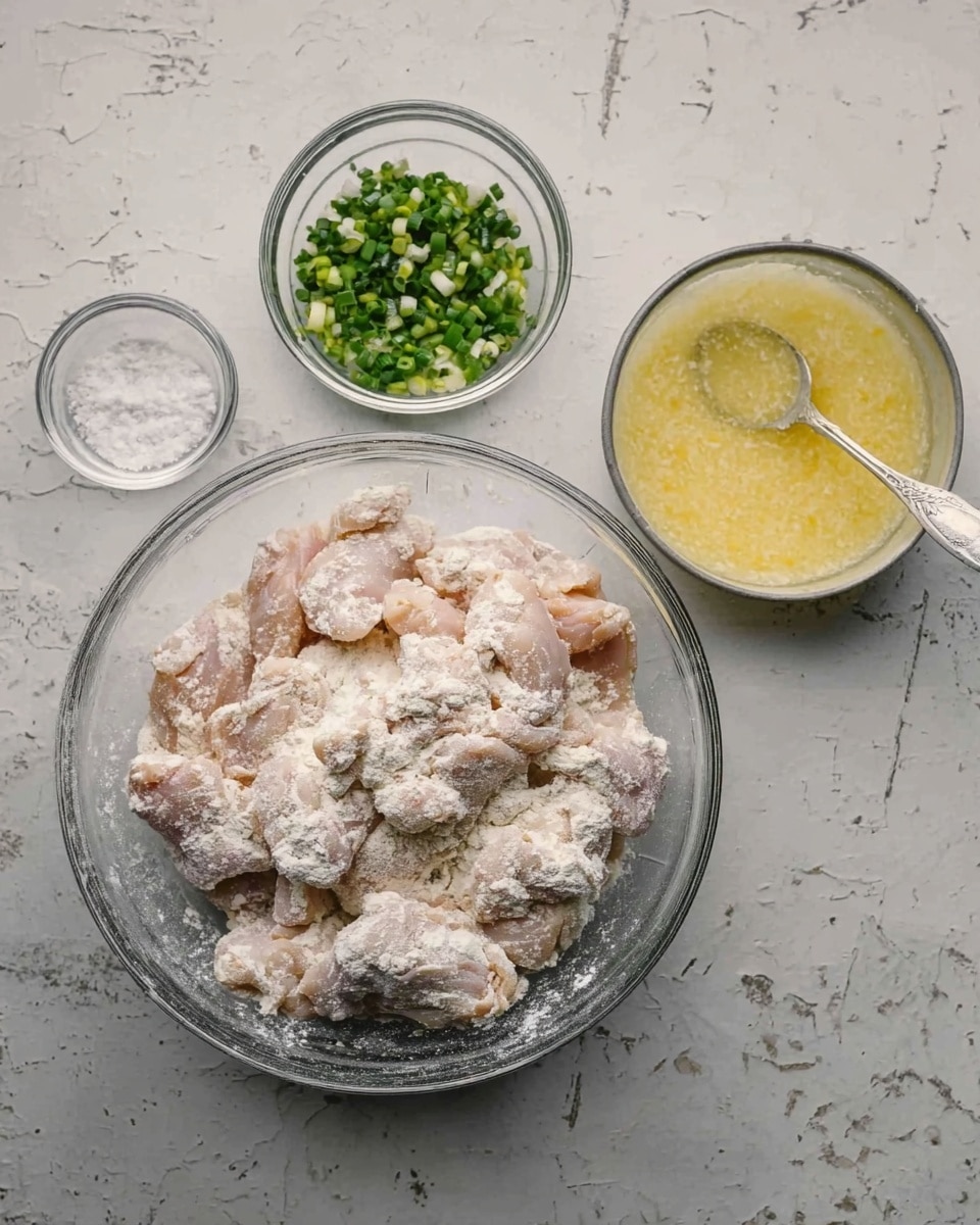 In the image, there is a large clear glass bowl in the center filled with raw chicken pieces covered with a layer of white flour, giving them a powdery look. To the left of the bowl, two smaller clear glass bowls hold finely chopped green onions and minced garlic. On the right side, another clear glass bowl contains a pale yellow liquid mixture with a spoon inside, likely an egg wash. All bowls sit on a white marbled textured surface. Photo taken with an iphone --ar 4:5 --v 7