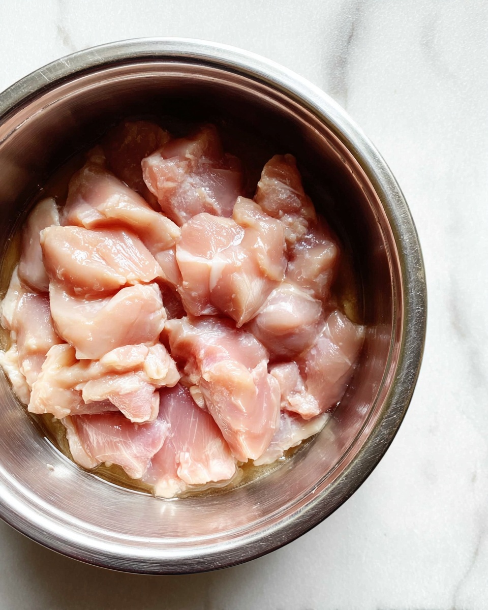 A close-up image of a silver metal bowl filled with raw, pale pink chicken pieces submerged in a little liquid, showing a mix of small and medium-sized chunks with a shiny, wet texture. The bowl is placed on a white marbled surface that adds a clean and simple background to the picture. The chicken pieces have a smooth, somewhat translucent look with light fat parts visible among them. Photo taken with an iphone --ar 4:5 --v 7