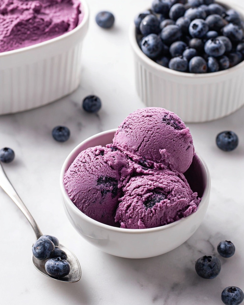 The image shows two scoops of purple-blueberry ice cream with a rough texture in a white bowl. Behind it, there is a second bowl filled with fresh blueberries, and to the left, a larger white container also has more of the same ice cream inside. Several blueberries are scattered around on a white marbled surface, with a silver spoon holding one blueberry placed near the bottom left corner. The overall scene is bright and clean with a focus on the vibrant color of the ice cream and blueberries. photo taken with an iphone --ar 4:5 --v 7