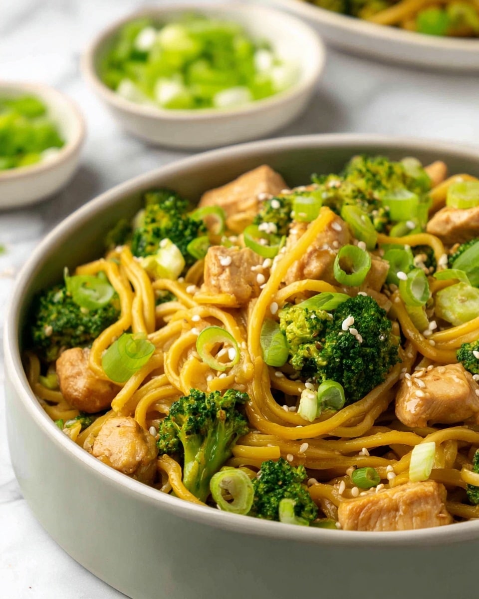 A close-up of a bowl with yellow noodles mixed with green broccoli pieces and bite-sized light brown chicken chunks. The noodles and chicken pieces are scattered evenly throughout, with small sliced green onions and white sesame seeds sprinkled on top. The bowl is white and set on a white marbled surface. In the background, there are small white bowls with sliced green onions and rice, blurred out slightly, adding depth to the image photo taken with an iphone --ar 4:5 --v 7