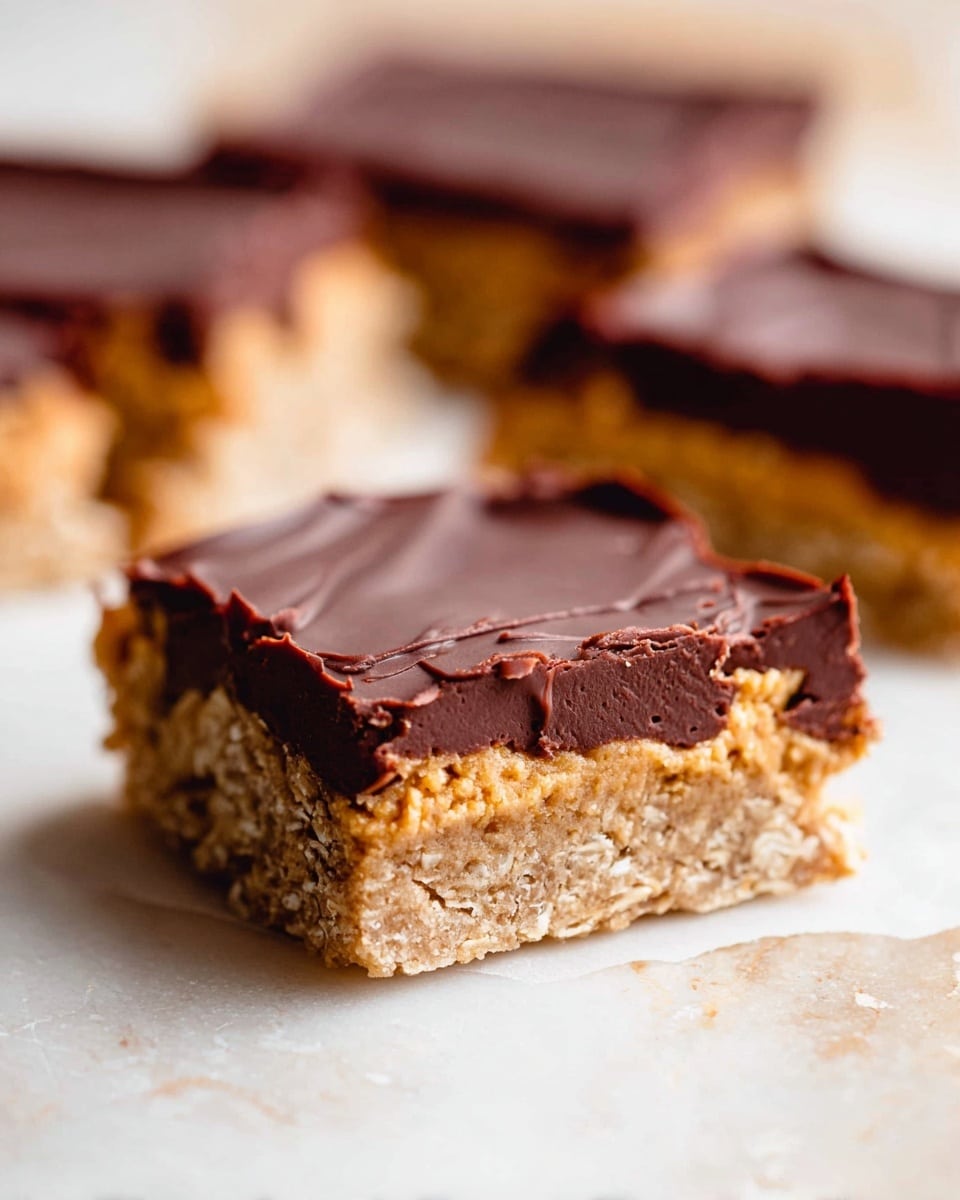The image shows a close-up of a square dessert bar with three distinct layers. The bottom layer is a light brown, crumbly oat base with visible oat flakes, giving a rough texture. The middle layer is a thick, glossy dark chocolate that looks smooth and firm. The top layer is a soft, golden cookie dough that looks slightly uneven with small lumps. The bar is placed on white marbled surface, and two more pieces of the dessert are blurred in the background. The focus is sharp on the front piece, showing the textures clearly. photo taken with an iphone --ar 4:5 --v 7