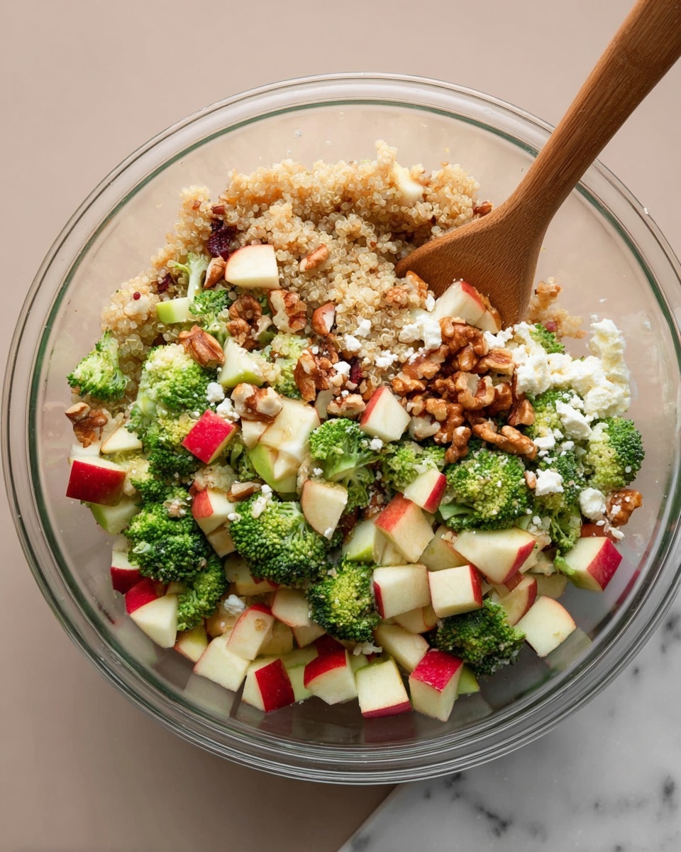 A clear glass bowl shows a colorful salad with a mix of many layers. At the base, there is light tan quinoa with a grainy texture. Scattered on top are green broccoli florets, small and fresh, adding a leafy feel. Small cubes of apple with white flesh and bright red skin are spread evenly, adding bright color. Light brown and tan chopped nuts, mainly walnuts and almonds, provide a crunchy texture throughout the salad. White crumbled cheese pieces are visible in bits, offering a soft contrast. A wooden spoon is partially inside the bowl, resting diagonally with its brown grain visible. The bowl is placed on a white marbled surface. Photo taken with an iphone --ar 4:5 --v 7