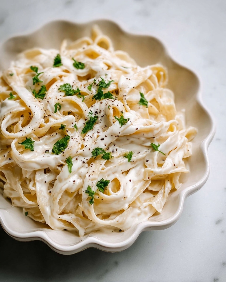 A white scalloped bowl filled with creamy white fettuccine pasta coated in a smooth sauce. The pasta strands are layered loosely with visible black pepper sprinkled on top. Small green parsley pieces are scattered over the pasta, adding a fresh touch. The bowl is placed on a white marbled surface. photo taken with an iphone --ar 4:5 --v 7