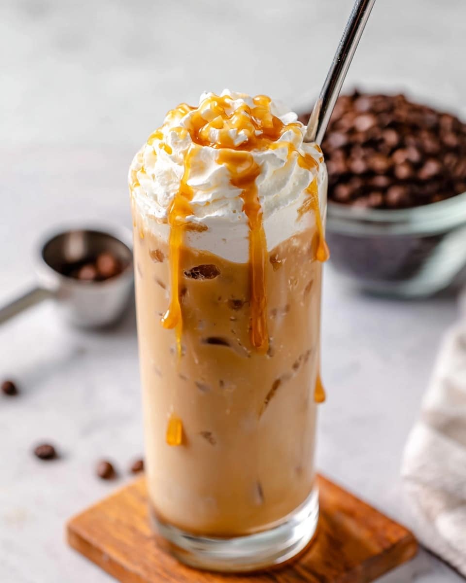 The image shows a tall clear glass filled with light brown iced coffee, with a few ice cubes visible inside. On top, there is a thick layer of white whipped cream, swirled and with a drizzle of golden caramel sauce dripping down the sides of the glass. The glass is placed on a small wooden board on a white marbled surface. In the background, there is a slightly blurred view of a clear bowl filled with dark brown coffee beans, with a silver scoop inside. Photo taken with an iphone --ar 4:5 --v 7