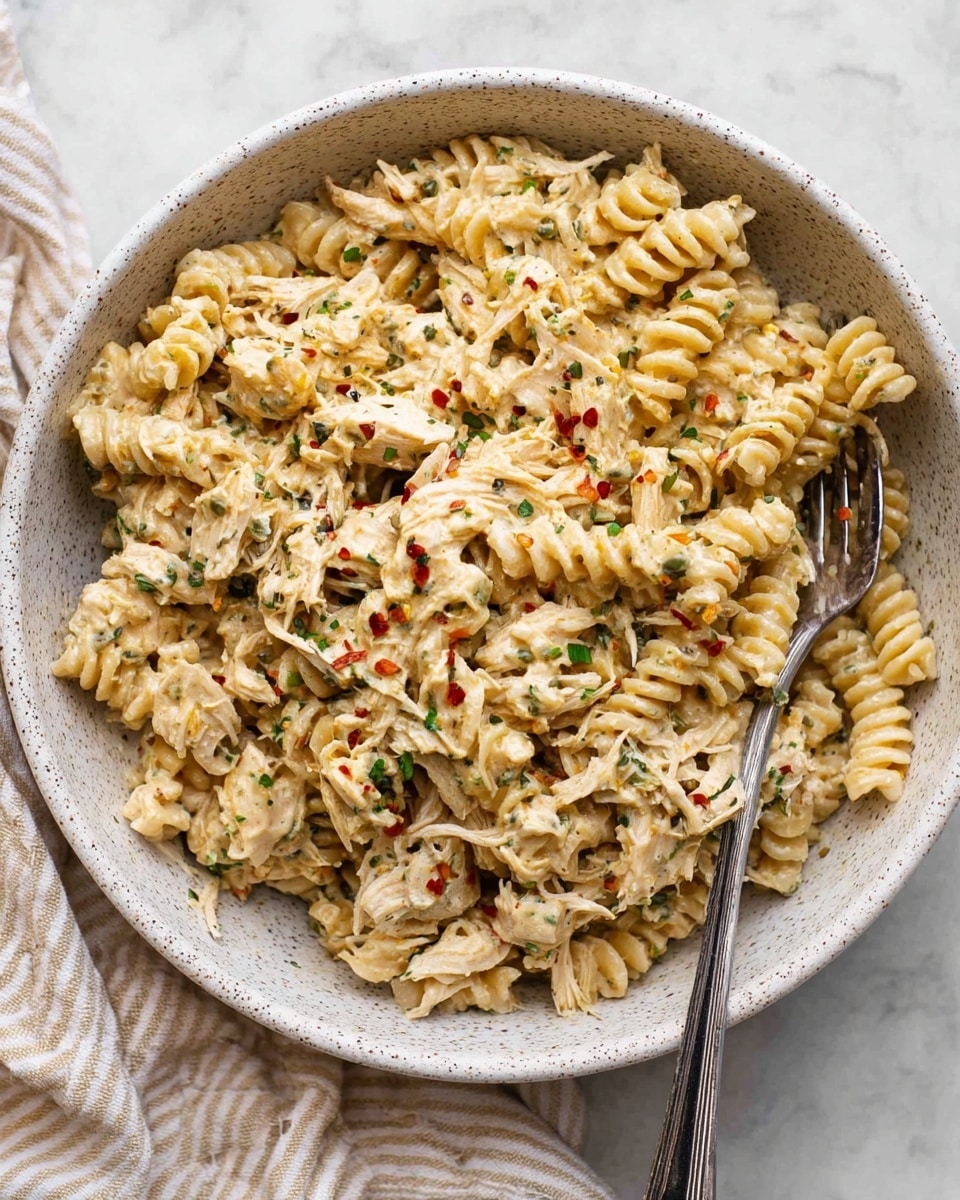 A bowl filled with creamy pasta layered evenly with spiral-shaped noodles coated in a light, creamy sauce mixed with shredded chicken pieces. The pasta and chicken are speckled with small bits of green herbs and tiny red pepper flakes, giving it a seasoned look. The dish is presented in a white speckled bowl, with a fork resting on the right side partially submerged in the food. The background shows a white marbled texture beneath the bowl, with a beige and white striped cloth peeking from the bottom left. Photo taken with an iphone --ar 4:5 --v 7