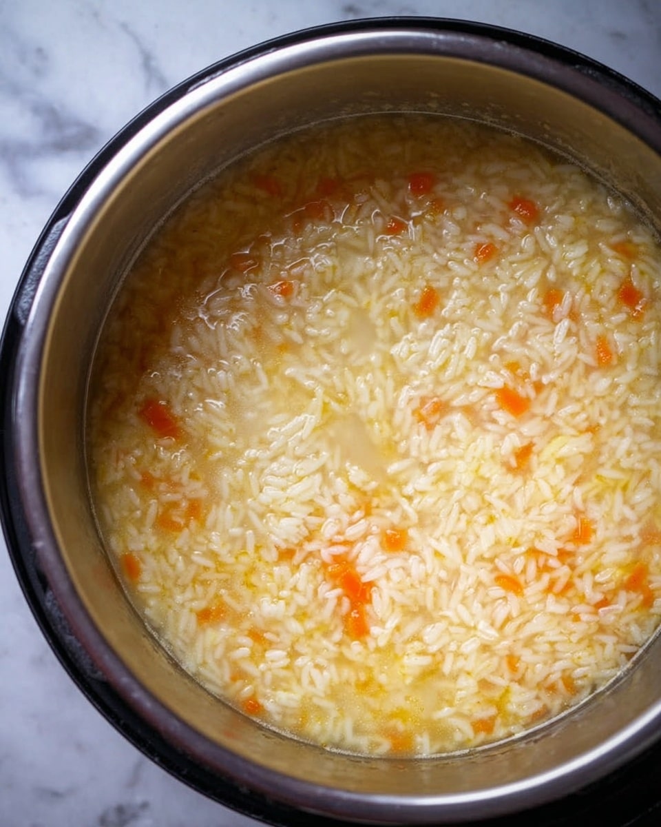 A close-up view of an open metal pot filled with cooked rice mixed with small orange carrot pieces. The rice is soft and sticky, with a light yellowish broth covering the grains, giving a moist texture. The pot is placed on a surface with a white marbled texture visible around the edges. photo taken with an iphone --ar 4:5 --v 7