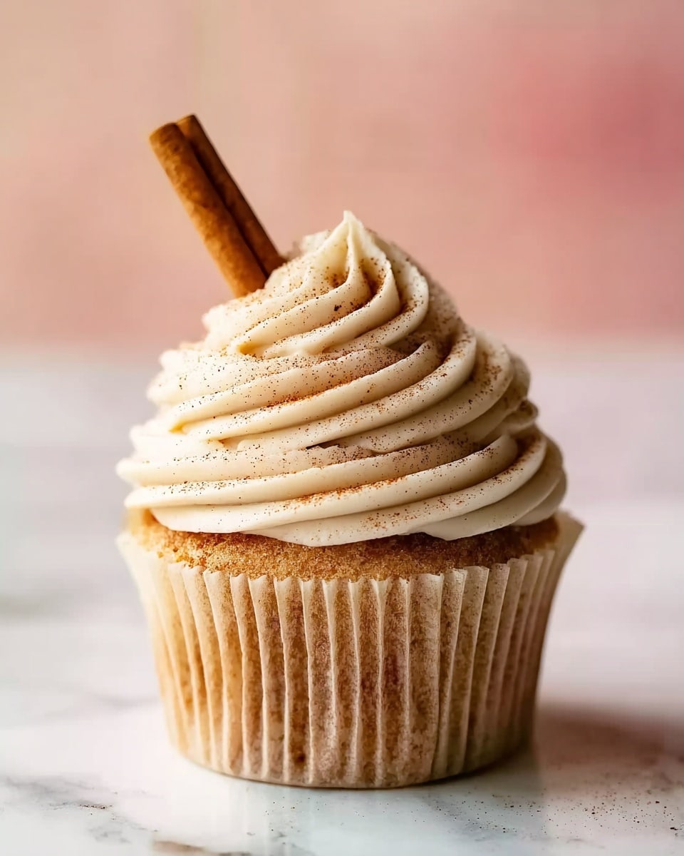The image shows a single cupcake with three visible layers: a soft light brown base in a beige paper liner, a tall swirl of creamy, off-white frosting with fine ridges covering the top, and a cinnamon stick inserted upright in the frosting along with a light sprinkle of cinnamon powder dusted over it. The cupcake is placed on a white marbled surface with a soft pink background. Photo taken with an iphone --ar 4:5 --v 7