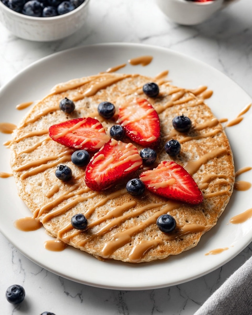 A single round pancake with a light brown, fluffy texture sits in the center of a white plate. On top, there is a drizzle of smooth, light brown sauce spread in thin lines across the whole pancake. Scattered on the pancake are fresh blueberries, small and dark blue, along with several bright red strawberry slices placed evenly, showing their inner soft texture and white seeds. The plate is set on a white marbled surface with a white bowl of extra blueberries blurred in the background. Photo taken with an iphone --ar 4:5 --v 7