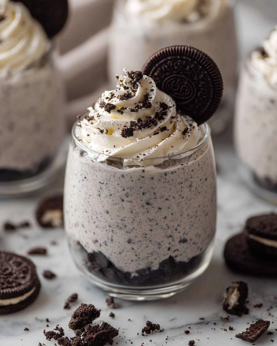 The image shows a clear glass filled with a light grey cream mixed with small dark cookie pieces, creating a speckled texture. At the bottom inside the glass, there is a layer of dark crushed cookies. On top, there is a swirl of white whipped cream with cookie crumbs sprinkled over it, and a whole dark chocolate cookie placed upright in the whipped cream. The glass is set on a white marbled surface scattered with whole and broken dark chocolate cookies. The blurred background shows more similar glasses filled with the same dessert. photo taken with an iphone --ar 4:5 --v 7