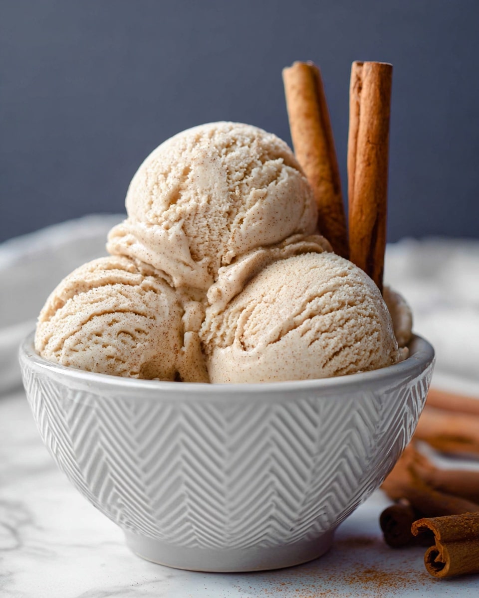 A white bowl with a textured chevron pattern holds four scoops of light beige ice cream with a creamy, slightly rough texture, closely packed together. Behind the ice cream, there are three upright cinnamon sticks with a rich brown color. The background is a white marbled surface with a soft gray backdrop. photo taken with an iphone --ar 4:5 --v 7