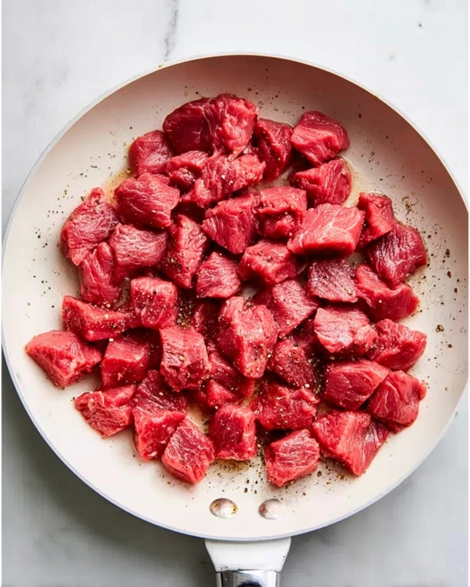 A white frying pan filled with many small pieces of raw red meat evenly spread inside it. The meat chunks have a fresh red color with some texture showing, and underneath them, the pan surface is visible in white with small bits of black pepper scattered around. The frying pan has a handle in the foreground, and the background is a white marbled surface. Photo taken with an iphone --ar 4:5 --v 7