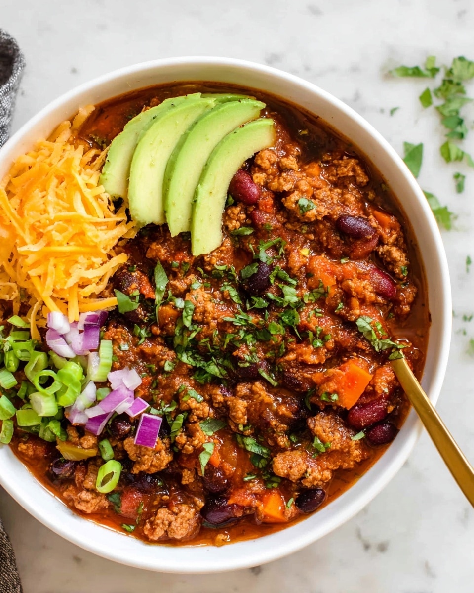 A white bowl filled with a thick chili made of cooked ground meat and red and black beans in a rich reddish-brown sauce, mixed with small pieces of bright orange carrot. On the left side of the bowl, there is a small pile of shredded orange cheese. Next to the cheese, there are three slices of pale green avocado, neatly layered. The top of the chili is sprinkled with chopped fresh green herbs and green onion slices, along with a few small pieces of purple onion. A golden spoon is partially visible on the right side of the bowl. The bowl sits on a white marbled surface. Photo taken with an iphone --ar 4:5 --v 7