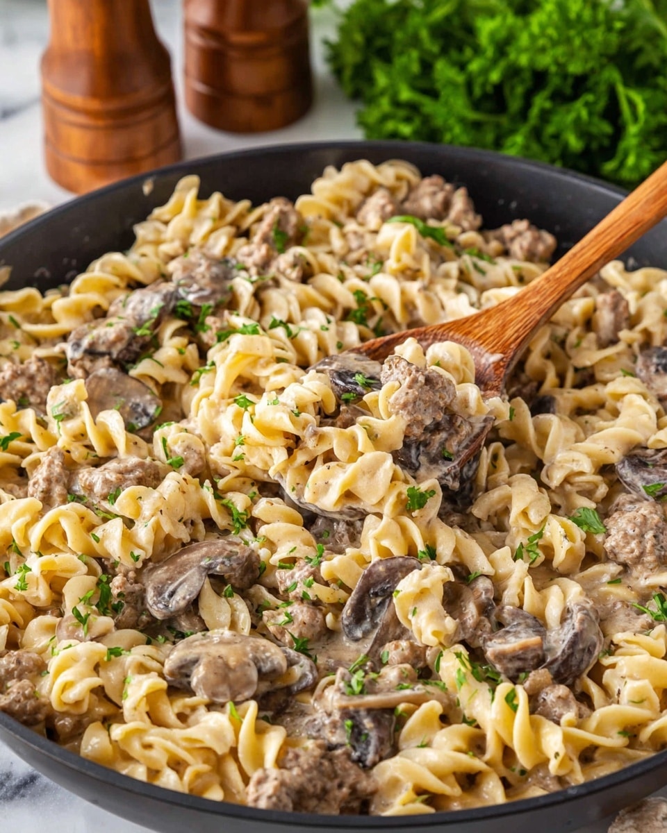 A close-up of a black skillet filled with creamy pasta made of curled, light yellow noodles mixed with small brown meat pieces and sliced dark brown mushrooms. The creamy sauce coats the pasta and meat evenly, with some bright green parsley sprinkled on top for color. A wooden spoon is stirring the pasta, lifting a small portion that shows the thickness of the noodles and sauce. The background shows a bunch of fresh green parsley and wooden salt and pepper shakers on a white marbled surface. photo taken with an iphone --ar 4:5 --v 7