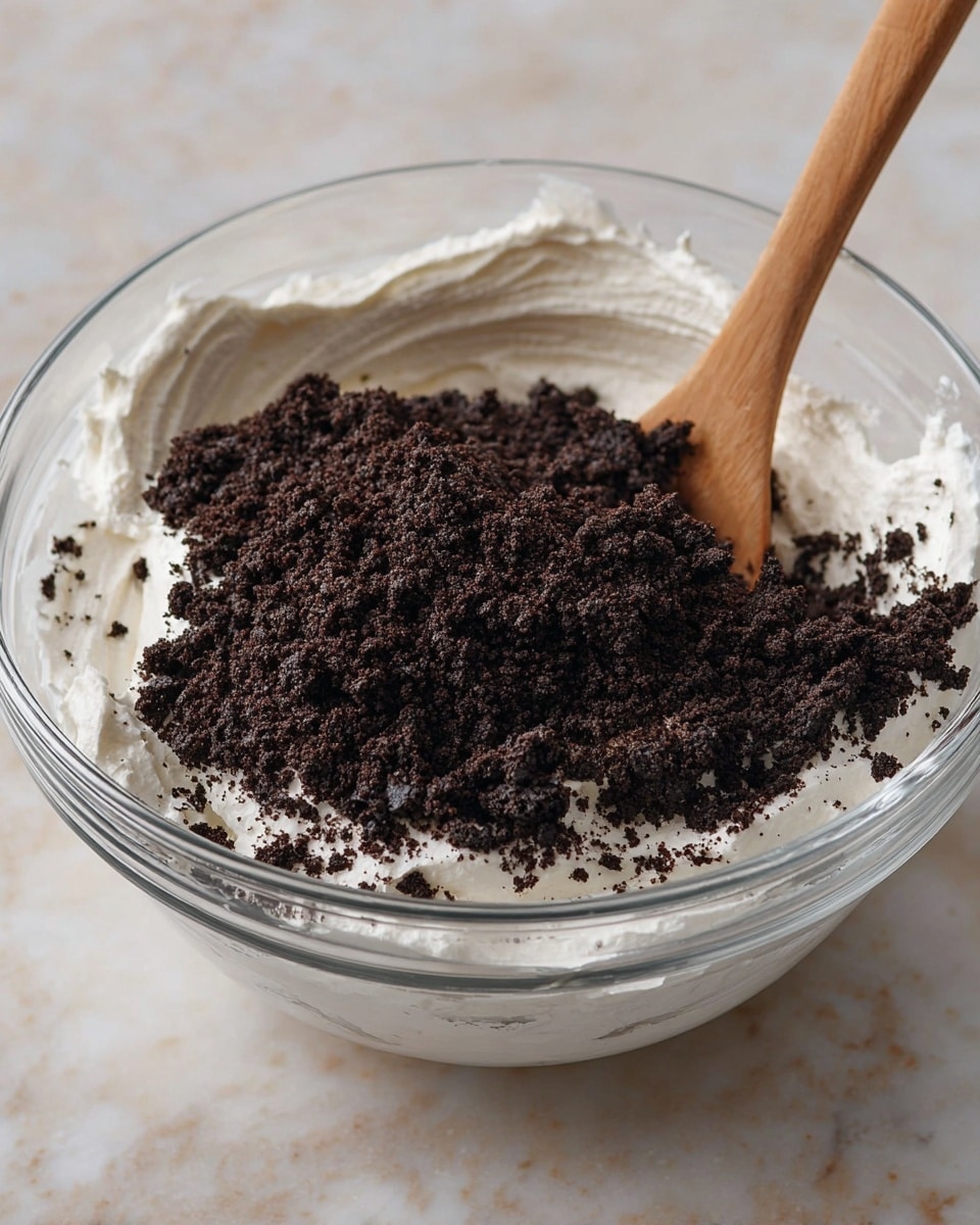 A clear glass bowl filled with two layers: the bottom layer is thick, white whipped cream with soft peaks and creamy texture, and the top layer is a generous heap of dark brown, coarsely crushed cookie crumbs spread unevenly. A wooden spoon is placed upright inside the bowl, with its handle extending out of view. The bowl sits on a white marbled surface. photo taken with an iphone --ar 4:5 --v 7