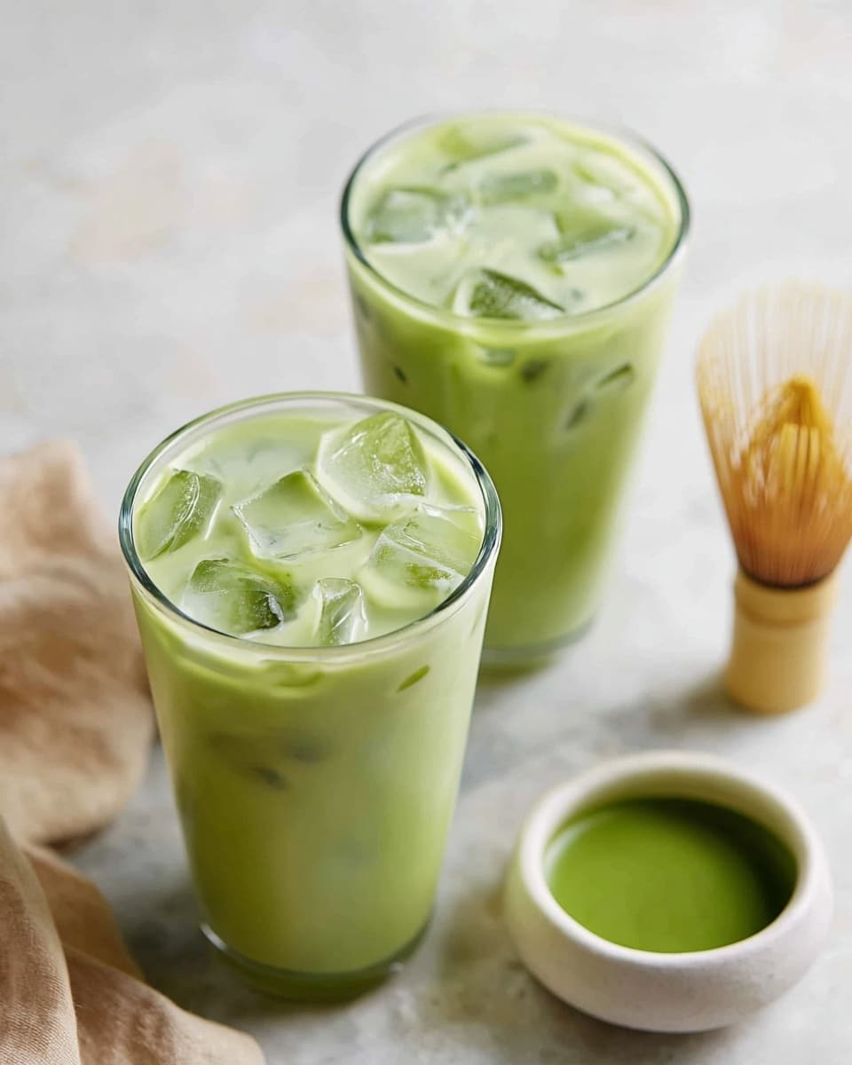 Two tall clear glasses filled with pale green iced matcha latte, each glass containing irregular ice cubes that float near the top. Behind the glasses is a small white bowl with a smooth green liquid inside, likely matcha tea. To the right, a bamboo matcha whisk lies on a white marbled surface, and to the left, a soft beige cloth is partially visible. The whole scene sits on a white marbled background, with soft natural light giving a fresh and clean look. Photo taken with an iphone --ar 4:5 --v 7
