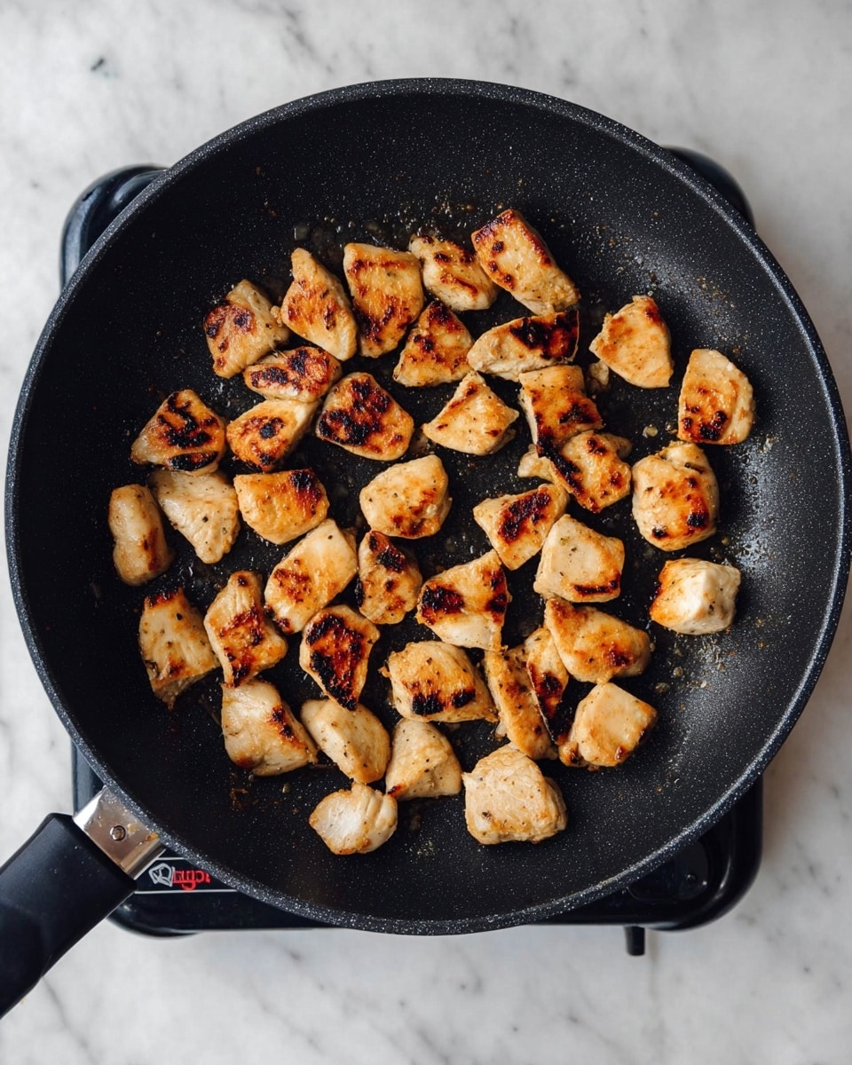 A black frying pan holds about 20 small pieces of cooked chicken with golden brown and slightly charred spots. The chicken pieces are uneven in shape and size, spread out in the pan on a black electric stove burner. The background is a white marbled texture. photo taken with an iphone --ar 4:5 --v 7