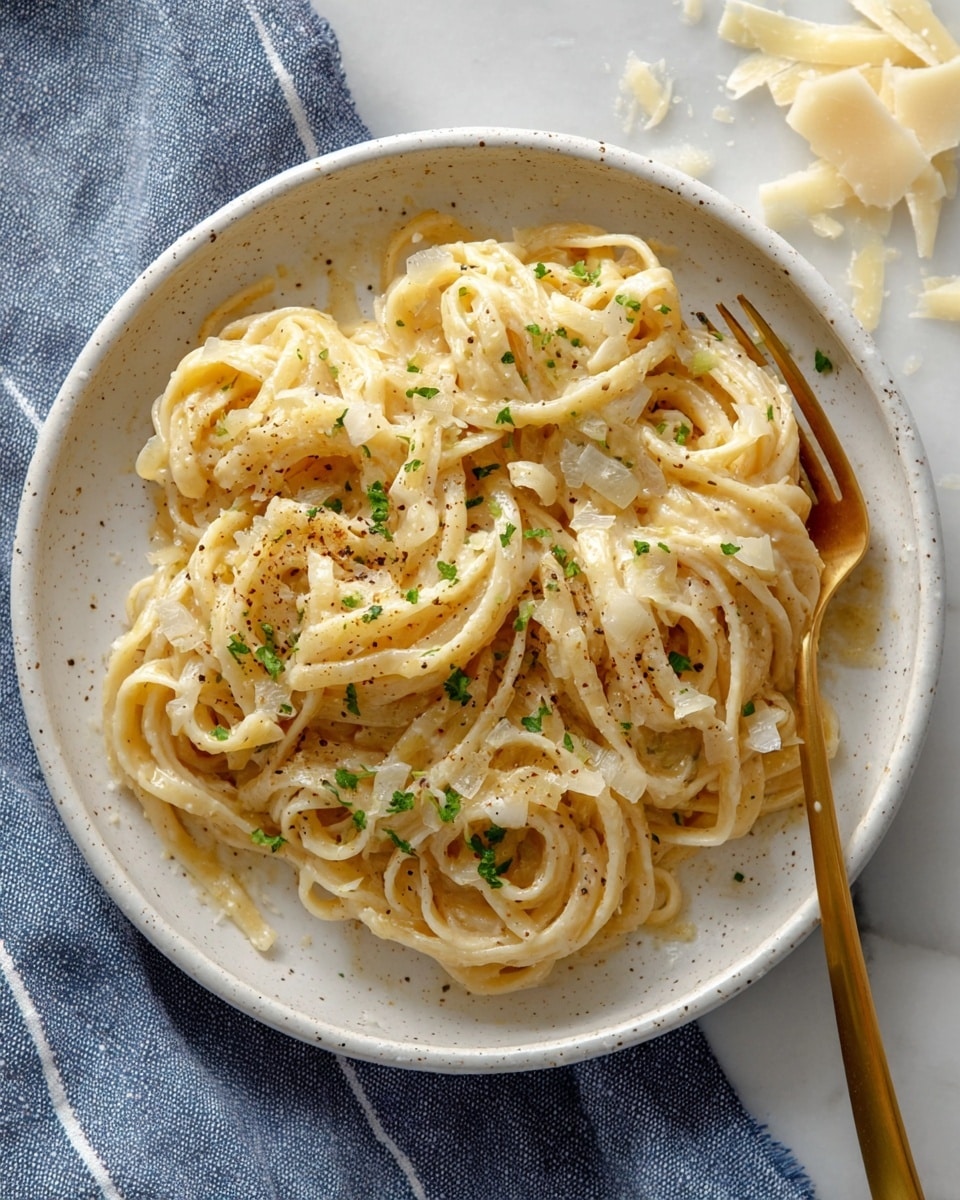 A white speckled bowl holds a serving of thick, pale yellow noodles mixed with small bits of sautéed onions and sprinkled with black pepper and chopped green herbs, arranged in a loose, tangled pile. A gold fork is partially inserted into the noodles on the right side, resting on the bowl’s edge. The bowl sits on a blue cloth with white stripes, placed on a white marbled surface. Small shavings of pale cheese are scattered on the upper right of the surface near the bowl. Photo taken with an iphone --ar 4:5 --v 7