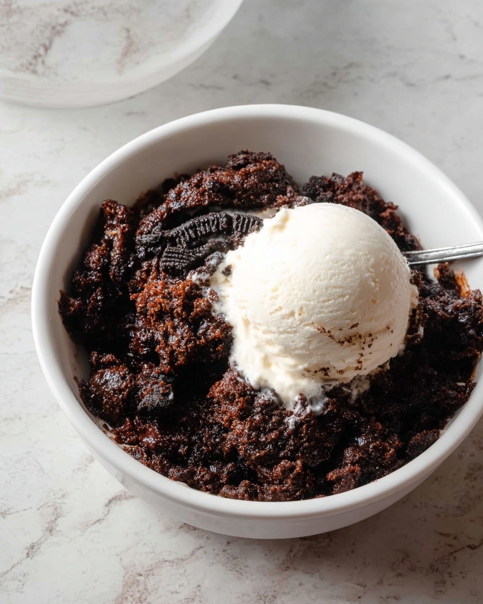 A white round bowl filled with a dark brown chunky chocolate base mixed with broken pieces of black chocolate cookies, topped with a smooth, round scoop of creamy white vanilla ice cream placed slightly off center; the texture of the chocolate base looks moist and dense with visible cookie bits. The bowl sits on a white marbled surface. photo taken with an iphone --ar 4:5 --v 7