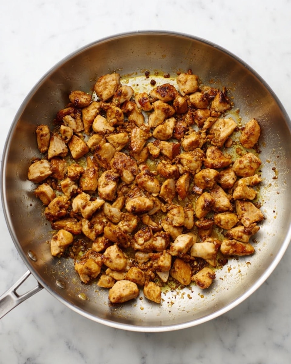 A stainless steel pan filled with small, browned pieces of cooked chicken spread evenly across the pan’s surface. The chicken pieces have a rich golden-brown color with some darker crispy edges, showing a textured, juicy look. The pan has some oil and browned bits that add a shiny and slightly glossy appearance. The background is a white marbled texture. photo taken with an iphone --ar 4:5 --v 7