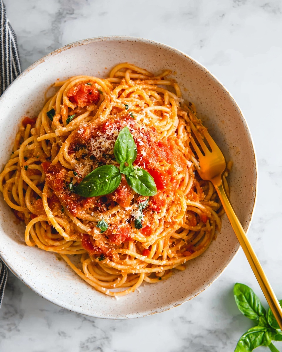 A white bowl filled with thick spaghetti layered at the bottom with long, twisted noodles in light orange color. On top, there is a chunky bright red tomato sauce spread unevenly across the noodles. A sprinkle of finely grated light yellow cheese is scattered over the sauce. In the center, three fresh green basil leaves add a pop of color. A gold fork and spoon rest on the right side with the fork partly buried in the pasta. The bowl sits on a white marbled surface with a small basil sprig on the right edge. Photo taken with an iphone --ar 4:5 --v 7