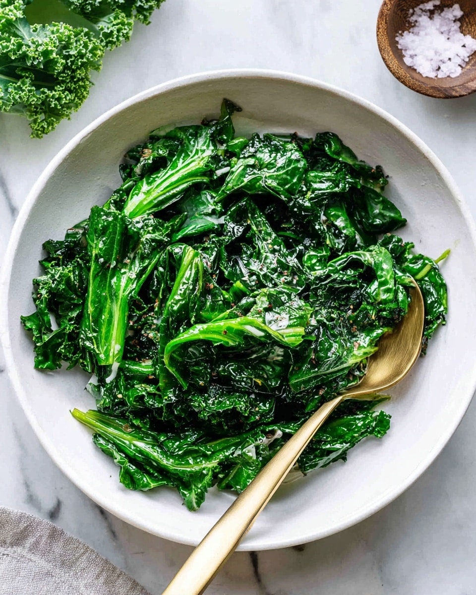 A white bowl is filled with cooked leafy greens that have a shiny, slightly wilted texture. The greens are bright and deep green with some visible stems and curly edges. A gold spoon rests inside the bowl, partially covered by the greens. The bowl sits on a white marbled surface, and some kale leaves and a small bowl of coarse salt are placed nearby. photo taken with an iphone --ar 4:5 --v 7