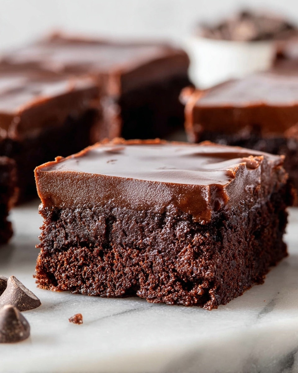 A close-up image of a chocolate brownie piece placed on a white marbled surface. The brownie has two visible layers: the bottom layer is thick, dense, and fudgy with a rich dark brown color and a slightly crumbly texture, and the top layer is a smooth, glossy chocolate frosting with a shiny dark brown finish. The edges of the brownie show a soft, moist interior, while the top has a silky, even spread. In the background, other similar brownie pieces are slightly out of focus, and small chocolate chunks are scattered on the white marbled surface in the front. Photo taken with an iphone --ar 4:5 --v 7