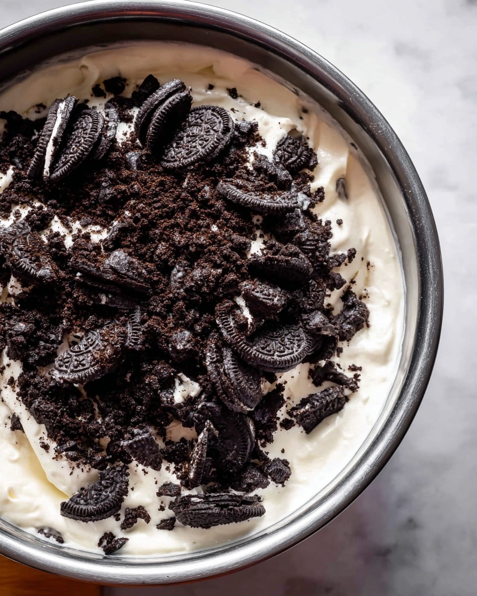 A close-up view of a metal bowl filled with a thick white cream layer at the bottom, topped with a generous pile of broken dark chocolate sandwich cookie pieces mixed with white cream filling. The bowl is on a white marbled surface providing soft contrast. photo taken with an iphone --ar 4:5 --v 7
