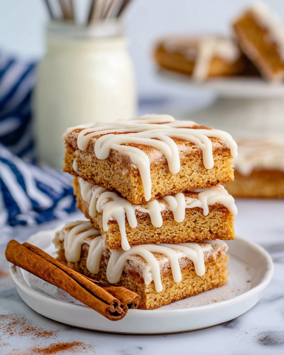 The image shows three stacked rectangular bars with a light brown, soft cake texture and a cinnamon sugar coating on top, sitting on a white plate on a white marbled surface. Each bar is covered with thick white icing drizzled in wavy lines that slightly drip down the sides. A cinnamon stick lies in front of the bars on the plate. In the background, there is a glass jar with white creamy frosting and a blurred blue and white striped cloth with forks. Photo taken with an iphone --ar 4:5 --v 7