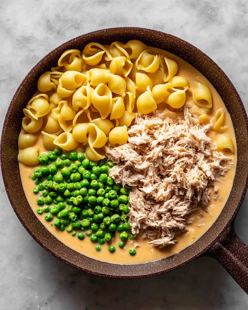 A dark brown pan filled with creamy light orange sauce as the base layer, topped with three distinct sections: bright yellow shell pasta on the right, fresh green peas on the upper left, and shredded light brown tuna on the lower left, all ingredients sitting separated on the sauce. The scene is set on a white marbled surface. photo taken with an iphone --ar 4:5 --v 7