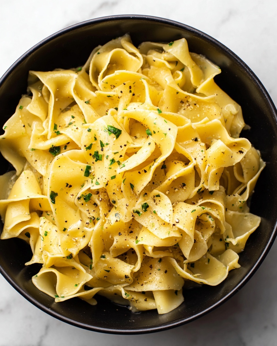 The image shows a black bowl filled with wide, soft yellow pasta pieces that have a gently twisted and folded shape. The pasta is lightly coated with a glossy, oily sauce and sprinkled with small green herb pieces and tiny black specks of ground pepper for color contrast. The bowl sits on a white marbled surface, adding a clean and bright background to highlight the warm tones of the pasta. photo taken with an iphone --ar 4:5 --v 7