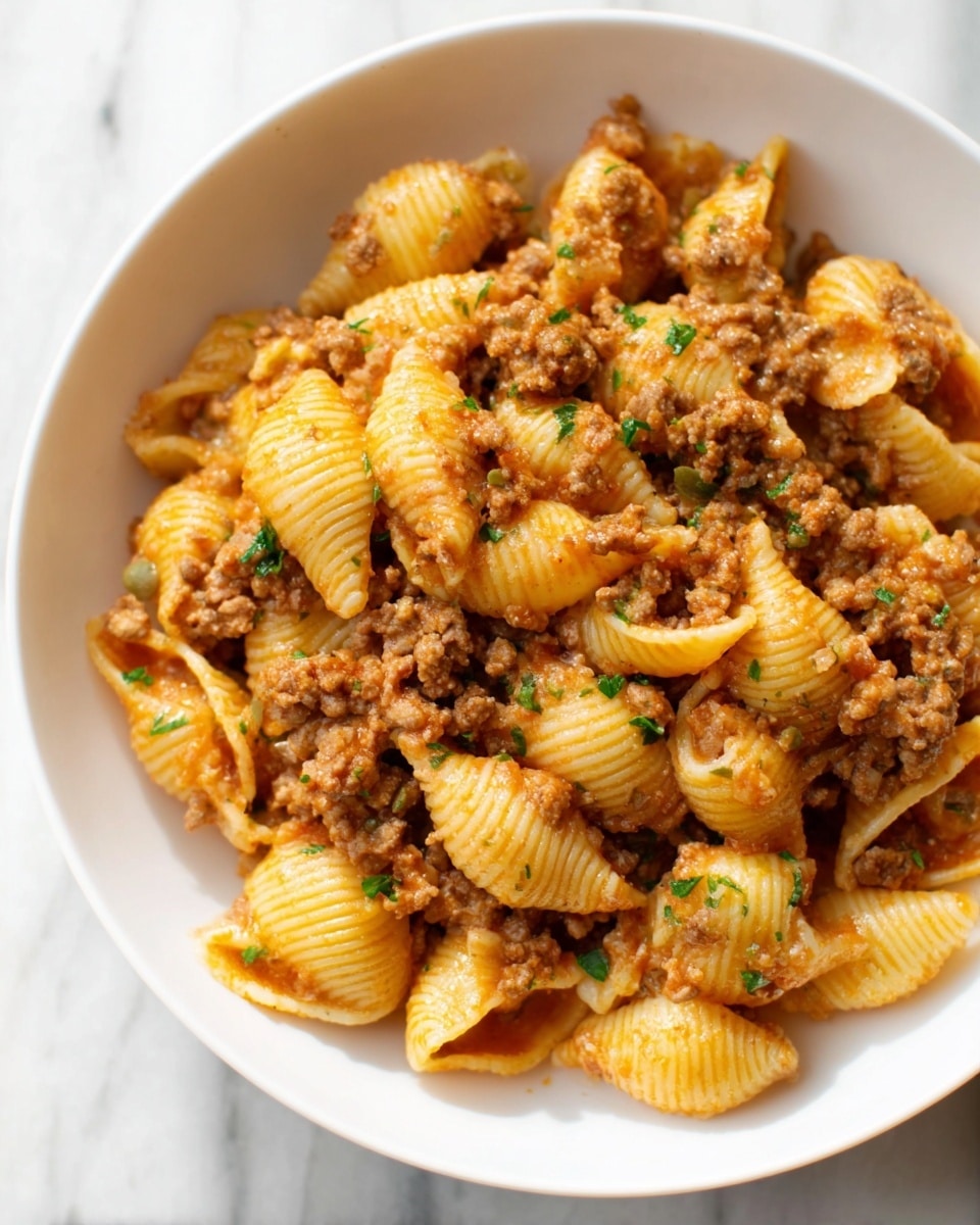 The image shows a close-up of a white bowl filled with pasta shells mixed with a minced meat sauce. The pasta shells are golden-yellow and have ridged textures. The meat sauce is brown with small onion pieces and coats the pasta evenly, blending into the crevices of the shells. Small green herb pieces are sprinkled on top, adding subtle color contrast. The bowl is placed on a white marbled surface. Photo taken with an iphone --ar 4:5 --v 7