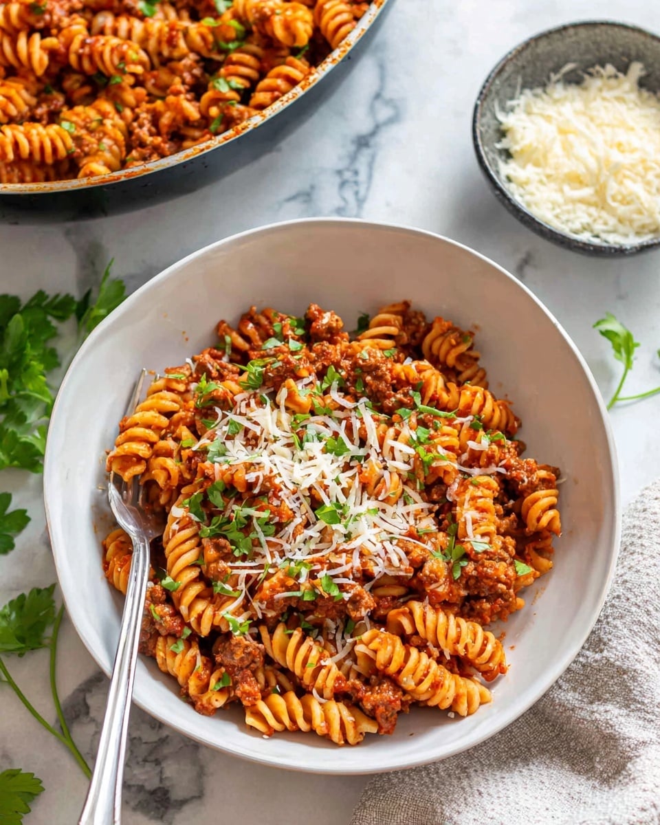 A white bowl filled with a layer of orange-brown spiral pasta mixed with minced meat and red tomato sauce, topped with white shredded cheese and scattered green herb leaves. A silver fork is stuck into the pasta on the left side of the bowl. In the background, there is a large pan of the same pasta dish and a small bowl with grated cheese, all set on a white marbled surface with some green herb sprigs nearby. A white cloth is partially visible on the right side. photo taken with an iphone --ar 4:5 --v 7