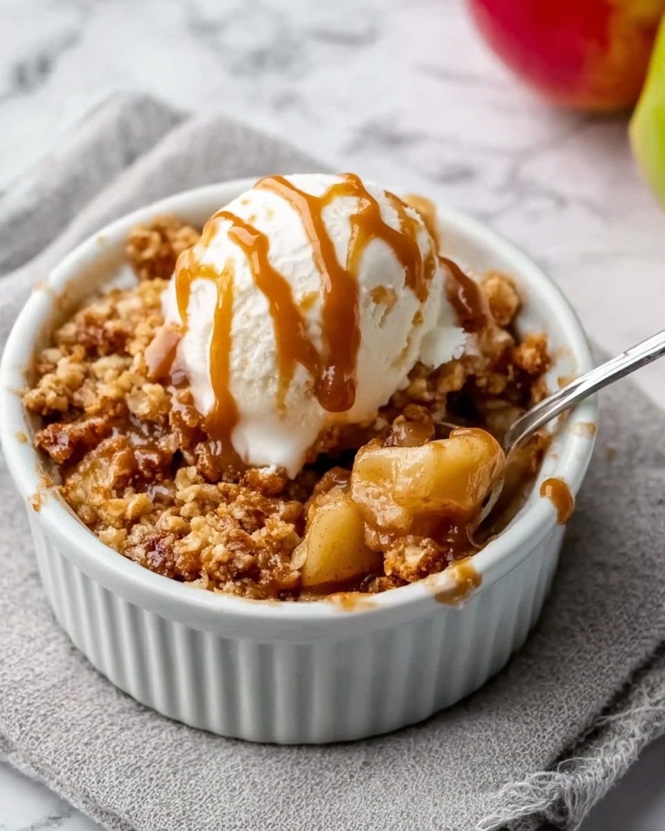 A white ramekin filled with warm apple crumble, showing soft, cooked apple pieces at the bottom covered by a golden brown crumbly topping. On top of the crumble is a scoop of creamy white ice cream drizzled with caramel sauce. A silver spoon is placed inside the ramekin, resting on the right side. The ramekin sits on a light grey cloth, with a white marbled surface in the background. The overall look is cozy and inviting, with the contrast of the warm dessert and cold ice cream clearly visible. Photo taken with an iphone --ar 4:5 --v 7