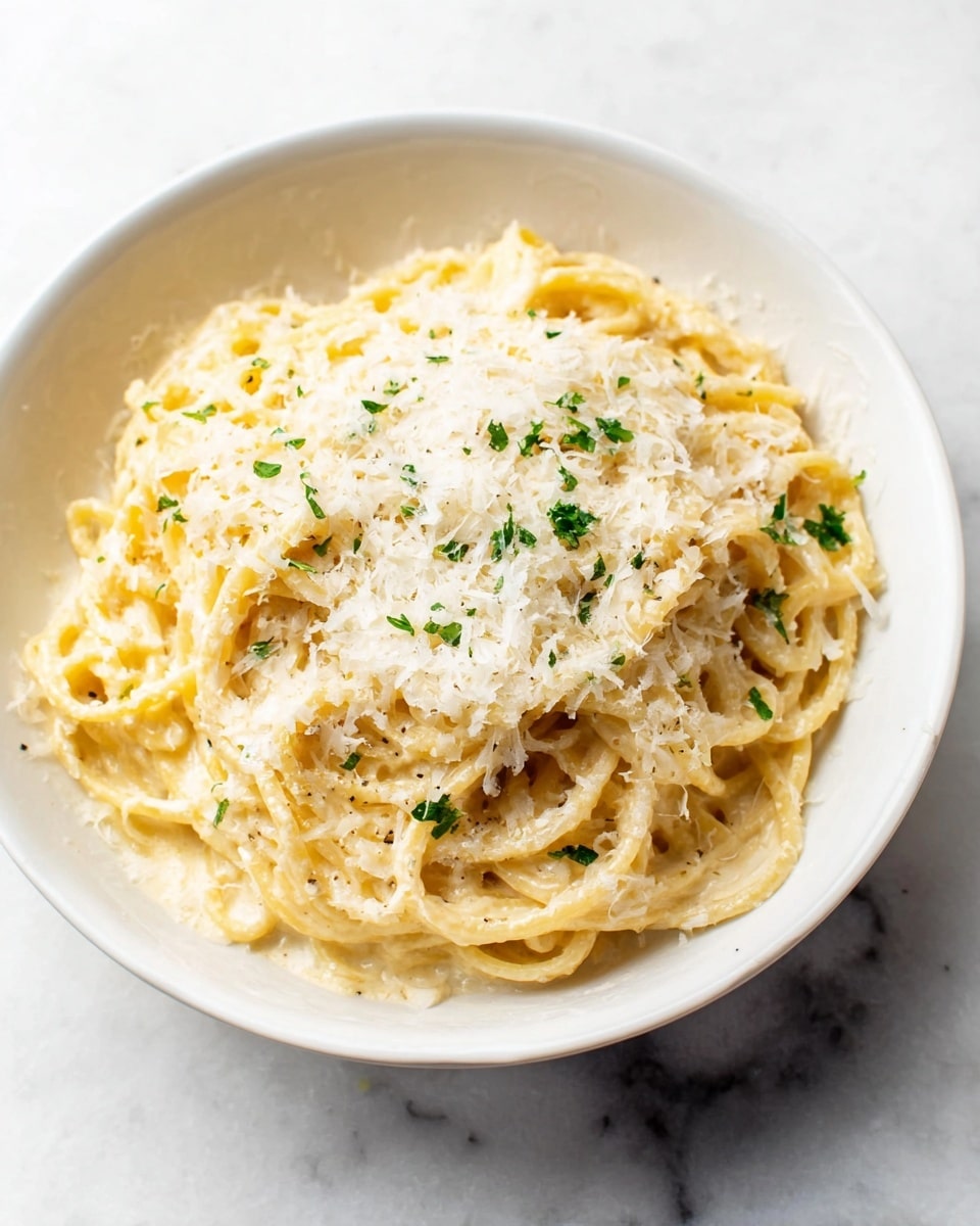 A white bowl holds a serving of creamy spaghetti pasta. The spaghetti noodles are coated in a smooth, pale yellow sauce and are loosely arranged in the bowl. On top, there is a thick layer of finely grated white cheese that covers most of the pasta. Small green herb pieces are sprinkled on the cheese for color contrast. The bowl is placed on a white marbled surface, adding texture to the plain background. photo taken with an iphone --ar 4:5 --v 7