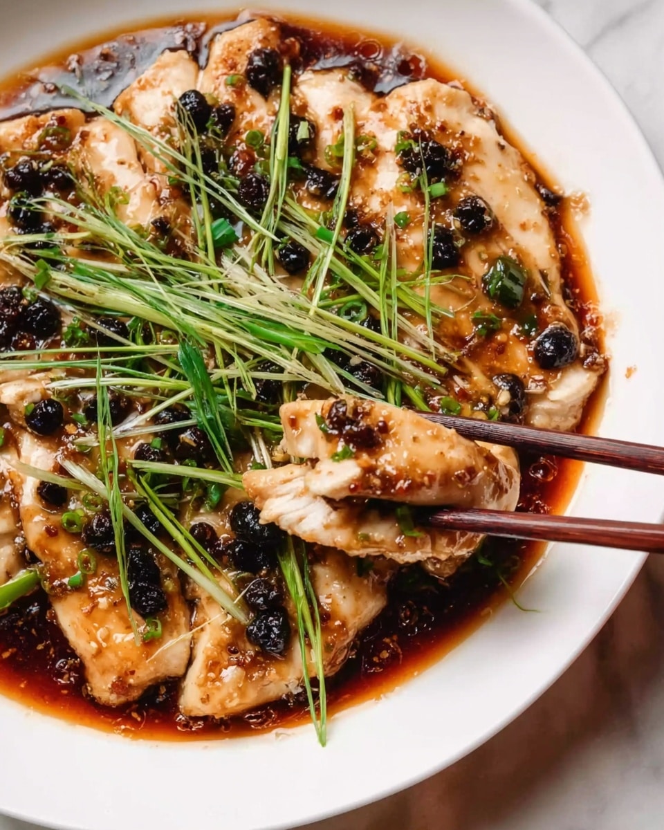 A white plate holds tender pieces of light golden cooked chicken covered in a shiny dark brown sauce. On top, there is a layer of thinly sliced green and white fresh scallions scattered all over. Small black fermented black beans are sprinkled throughout the dish, adding dark spots. A woman's hand holding wooden chopsticks is lifting one piece of chicken. The surface beneath the plate is a white marbled texture. photo taken with an iphone --ar 4:5 --v 7