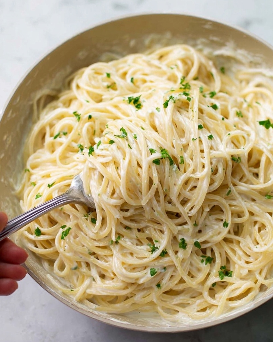 This image shows a close-up of creamy spaghetti pasta mixed with a white sauce. The spaghetti strands are coated evenly with the smooth, light-colored sauce, and small bits of green herbs are sprinkled on top, adding a fresh touch. A woman's hand is using a fork to lift some pasta from the white marbled surface beneath. The texture looks soft and creamy, with the pasta twisted around the fork and shining slightly from the sauce. The whole scene is bright and clean. photo taken with an iphone --ar 4:5 --v 7