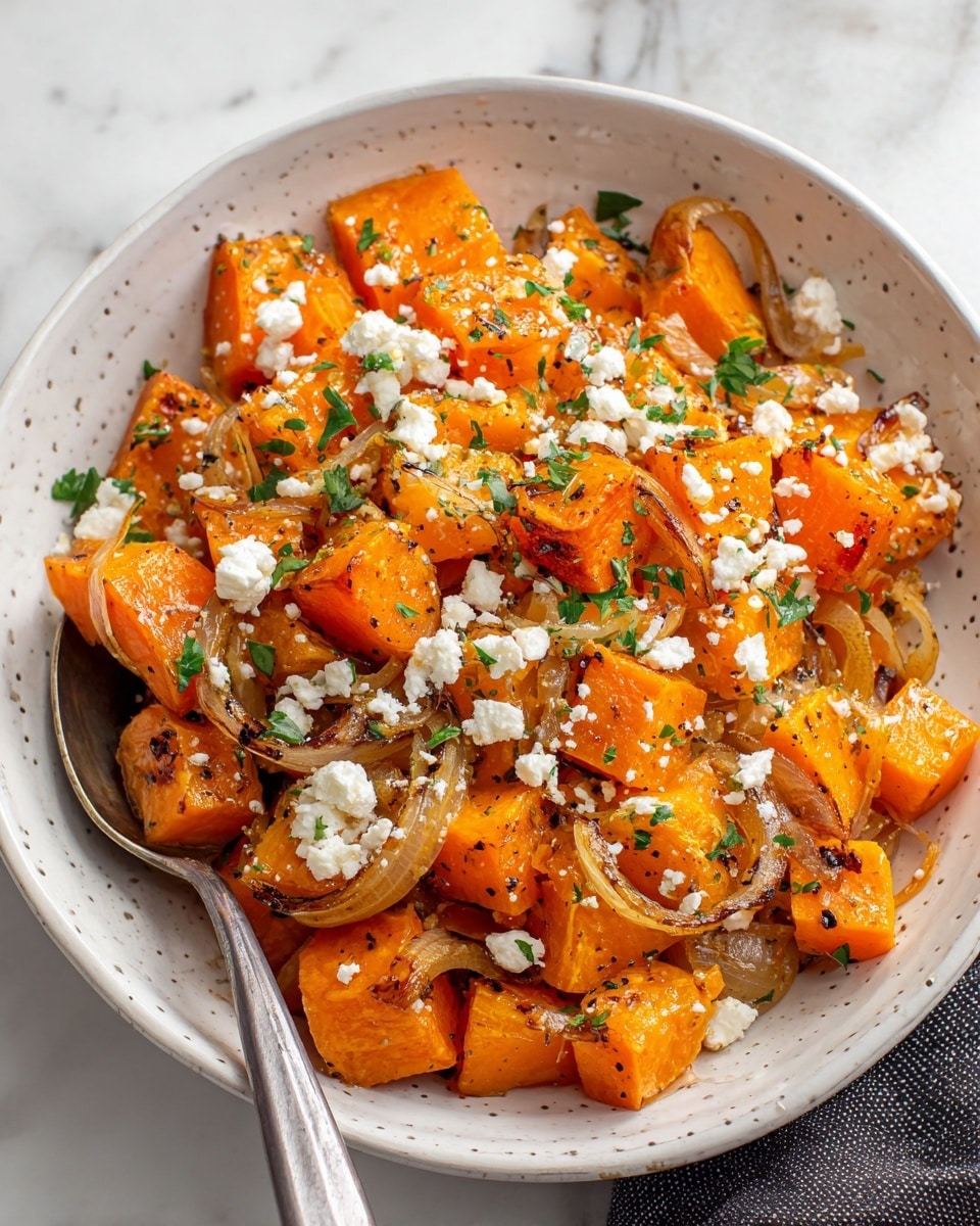 The dish shows roasted orange sweet potato chunks mixed with translucent sautéed onion slices in a white bowl with small raised dots on the inside. White crumbled cheese is sprinkled on top along with small green parsley leaves, adding a bit of fresh color. The sweet potatoes have a slightly browned texture, and the onions look soft and glossy. A silver spoon is partially visible resting inside the bowl on a white marbled surface. photo taken with an iphone --ar 4:5 --v 7