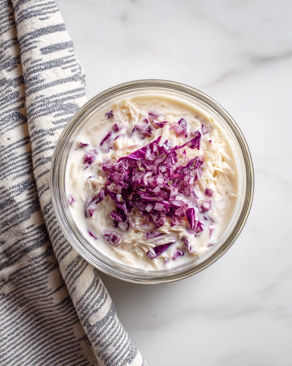 A clear glass bowl sits on a white marbled surface, filled with a light beige shredded mixture as the bottom layer. On top of this is a layer of white creamy sauce, slightly thick in texture. Small, finely chopped pieces of purple-red onion rest in the center of the bowl, adding a pop of color. The bowl is positioned near a striped gray and white cloth on the left side of the image. photo taken with an iphone --ar 4:5 --v 7