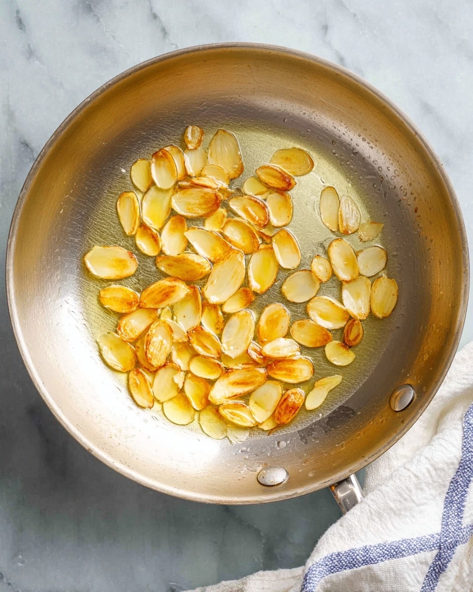 A shiny metal pan holds a single layer of thin, golden-brown garlic slices frying in clear oil. The garlic slices have a crispy texture and are scattered evenly within the warm oil. The pan sits on a white marbled surface, with a corner of a white cloth with blue stripes visible at the bottom right. The image is bright and focused from above, showing the details of the garlic and oil. photo taken with an iphone --ar 4:5 --v 7