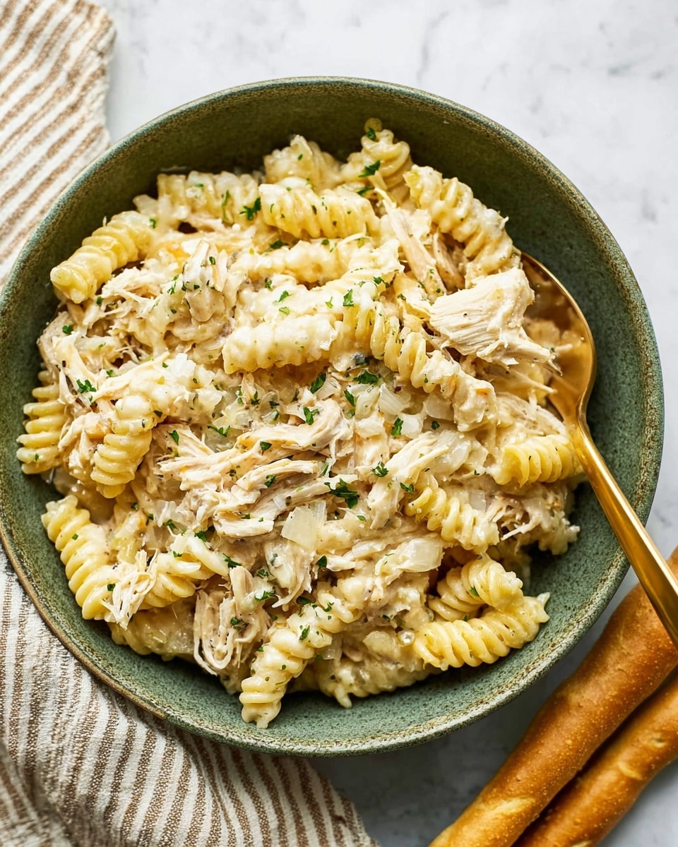A close-up view of a green bowl filled with a creamy pasta dish, showing two layers: a base of golden-yellow spiral pasta mixed with shredded light-colored chicken pieces, all coated in a smooth, white sauce with a slightly melted texture and flecks of green herbs sprinkled on top. A gold spoon is partially inside the bowl, resting on the pasta. Outside the bowl, on a white marbled surface, there are two light brown breadsticks placed near the bowl and a beige striped cloth underneath part of the bowl. photo taken with an iphone --ar 4:5 --v 7