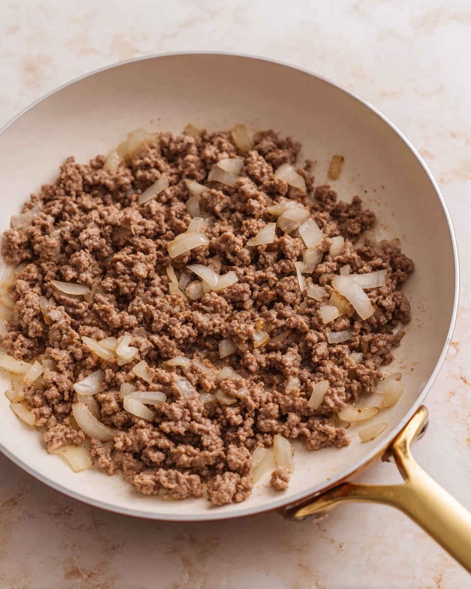 A white frying pan with a gold handle holds cooked ground meat mixed with small pieces of cooked onion. The meat is a brown color with a crumbly texture, and the onion pieces are translucent white with a soft look. The pan is on a white marbled textured surface, showing the cooked mixture evenly spread inside the pan photo taken with an iphone --ar 4:5 --v 7