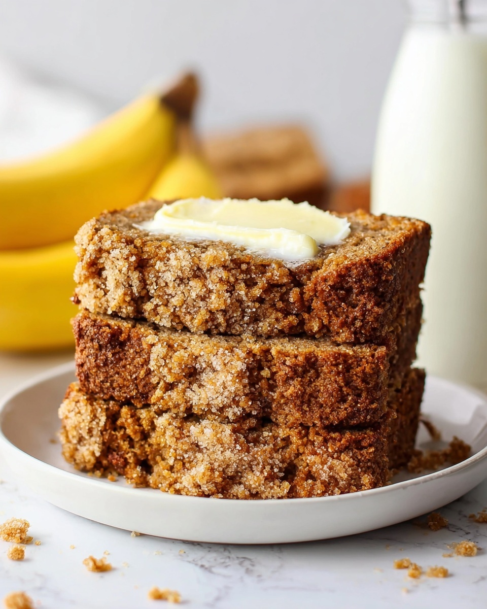 A white plate holds three thick slices of crumbly banana bread stacked on top of each other. The top slice is spread with slightly melted butter, showing a creamy white layer with soft texture contrasting the rough, golden-brown crust of the bread. The bread itself has a coarse, crumbly look with sugar crystals visible on the surface. In the background, there are three whole yellow bananas and a glass milk bottle placed on a white marbled surface. The setting is bright and clean, focusing on the warm, inviting texture of the banana bread. photo taken with an iphone --ar 4:5 --v 7