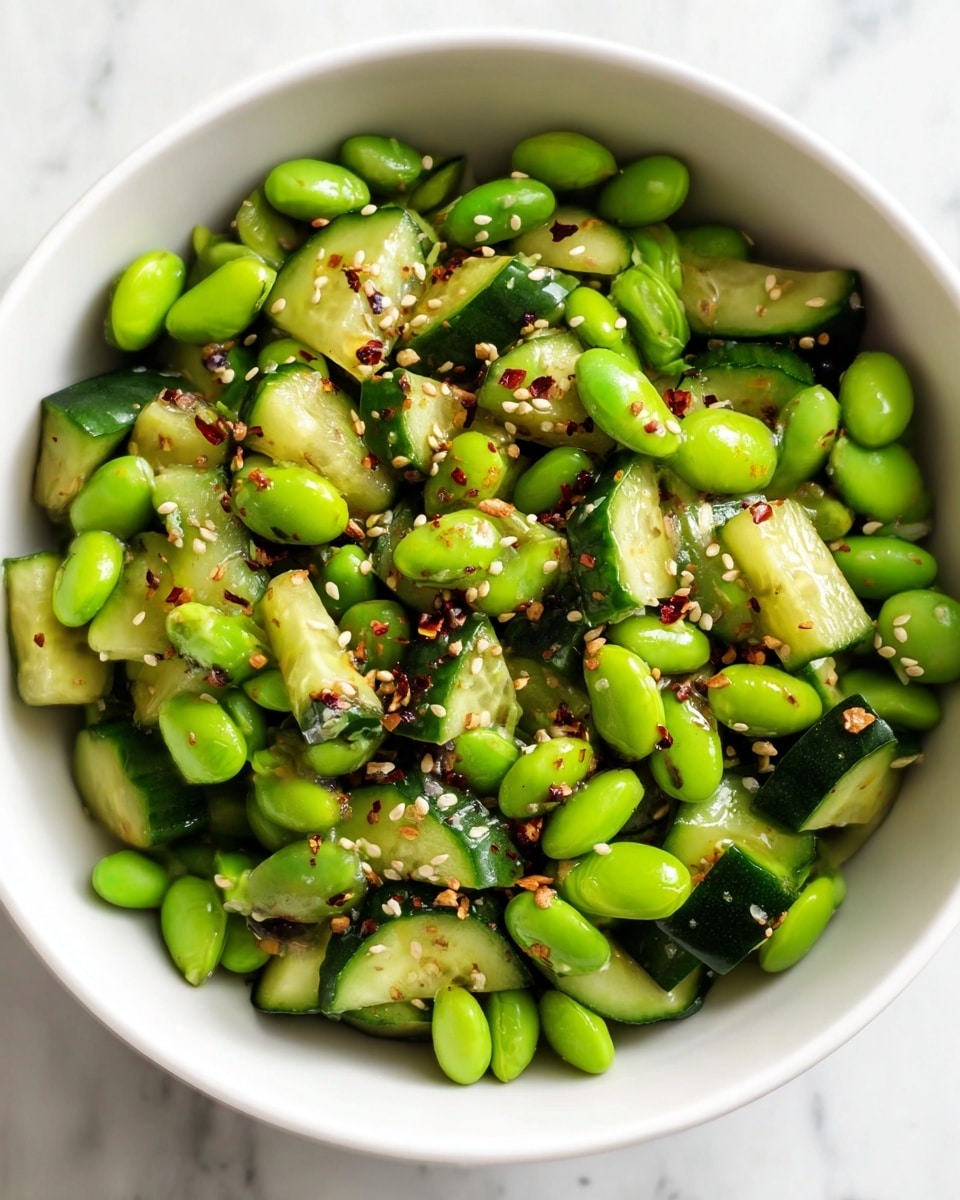 A white bowl filled with bright green edamame beans mixed with dark green cucumber pieces cut into bite-sized chunks. The salad is sprinkled with small white sesame seeds and red chili flakes evenly spread across the top, giving a contrast of colors. The edamame beans have a smooth, shiny texture, while the cucumber pieces show a mix of smooth flesh and a darker green ridged skin. The bowl sits on a white marbled surface, making the bright colors of the salad stand out well. photo taken with an iphone --ar 4:5 --v 7