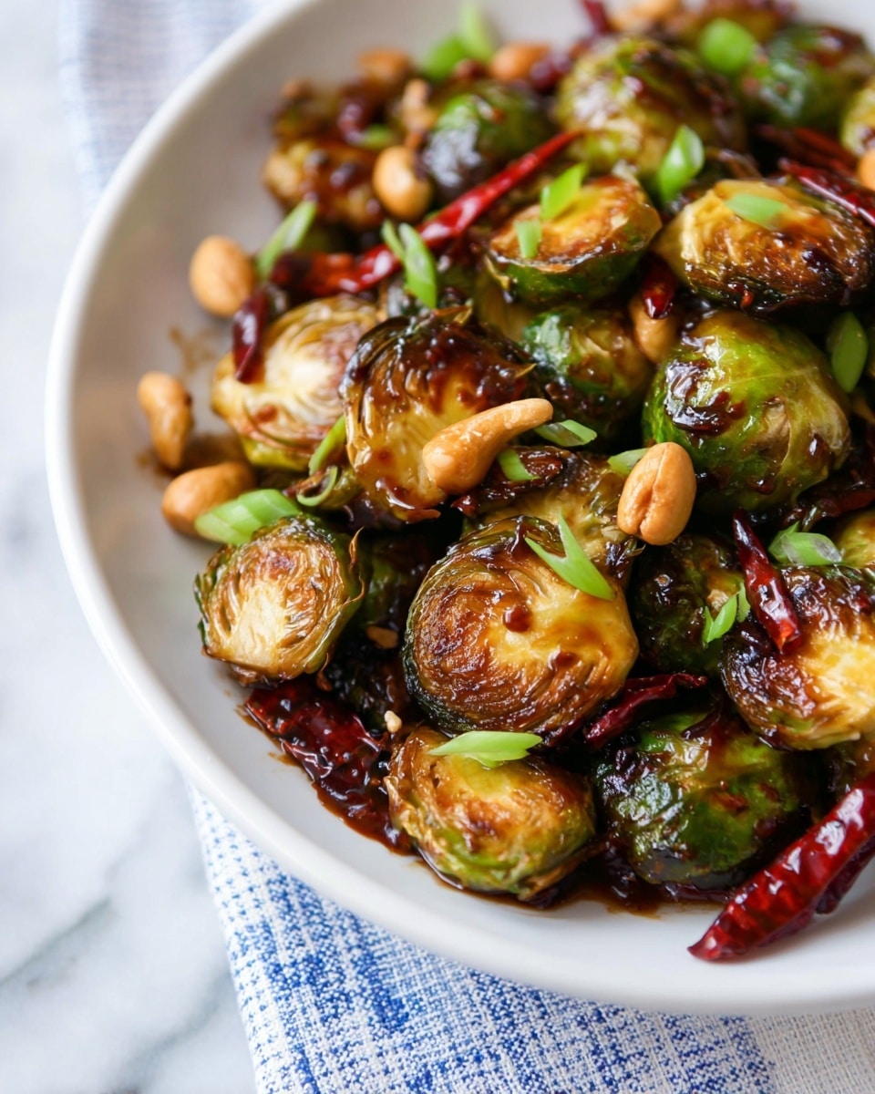 A close-up shot of one layer of roasted Brussels sprouts in a glossy, dark brown sauce, arranged densely in a shallow white plate. The Brussels sprouts are mostly halved and caramelized with a slight char, showing rich green and golden tones. Scattered throughout are small, light tan cashews and bright green chopped scallions, along with a few whole dried red chilies adding a deep red contrast. The plate sits on a white marbled surface with a soft blue checkered cloth partially visible underneath. The lighting highlights the shiny texture of the sauce and fresh ingredients, creating a vibrant and appetizing look photo taken with an iphone --ar 4:5 --v 7