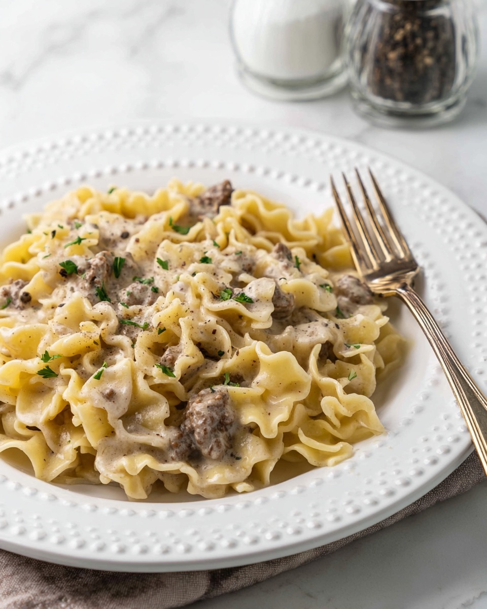 A close-up of creamy pasta served on a white plate with a raised dotted edge. The pasta is a light yellow color with ruffled edges and mixed with small pieces of browned meat. The creamy sauce is light beige and coats the pasta evenly. Small bits of green herbs are sprinkled on top, adding a fresh touch. There is a fork resting on the plate’s edge. The plate sits on a white marbled surface with a salt and pepper shaker blurred in the background. photo taken with an iphone --ar 4:5 --v 7